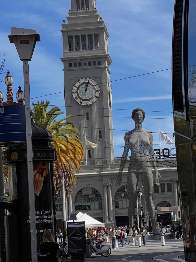 A large, translucent nude sculpture of a woman stands in front of a clock tower in San Francisco.